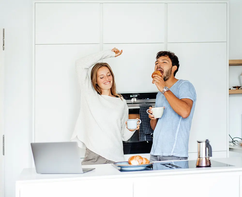 Junges Paar genießt Kaffee und Croissants in moderner Küche, fröhliche Stimmung am Morgen mit heller Kücheneinrichtung und Frühstücksszene.